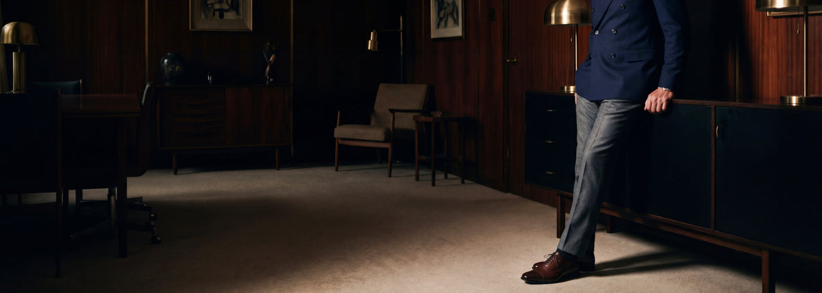 A man in a navy blazer, gray jeans, and men's brown Italian leather dress loafer shoes stands in a dimly lit, elegant room with wood-paneled walls and dark furniture.